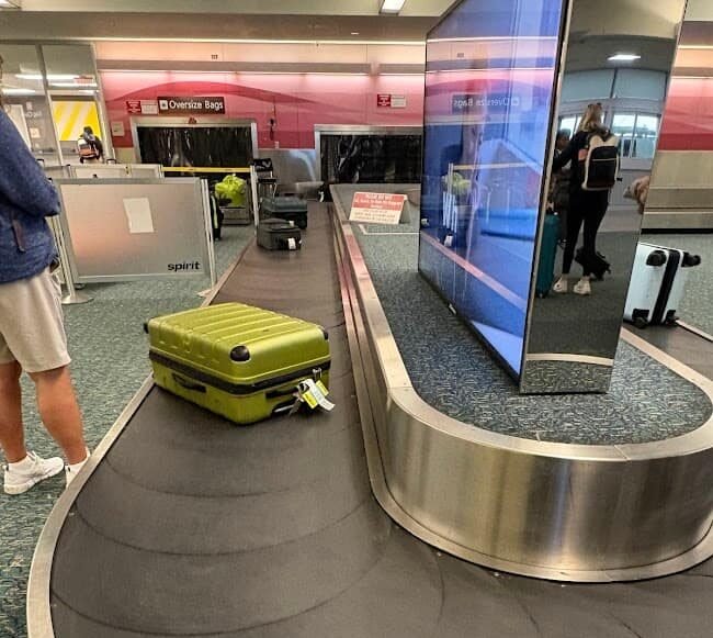 A lime green suitcase on a moving baggage carousel at an airport. A Spirit Airlines sign is visible in the background, showing travelers how to fly to Florida on a budget and collect their luggage at MCO.