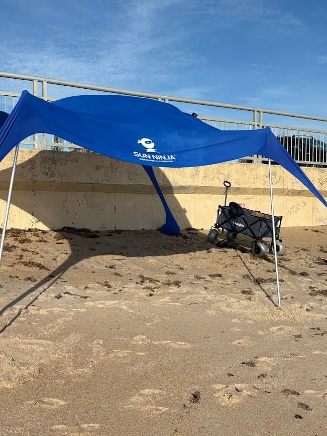 A blue Sun Ninja beach tent set up on the sand at New Smyrna Beach, providing shade next to a small beach wagon.