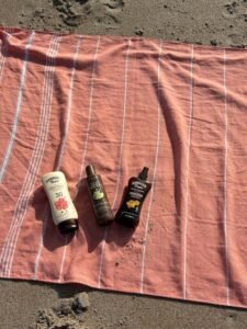 Three bottles of sunscreen, including Hawaiian Tropic SPF 30, resting on a pink striped BAY LAUREL Turkish beach towel on the sand.