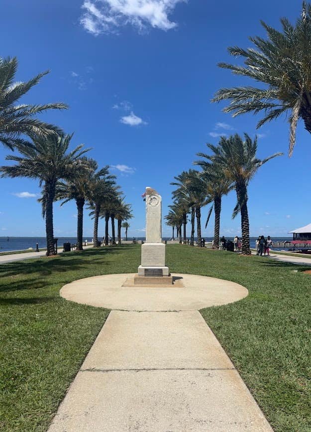A sunny view of a stone monument at Veterans Memorial Park in Sanford, Florida. Palm trees line a walkway leading toward the water, showing a relaxing spot to visit when you fly to Florida on a budget through Sanford.