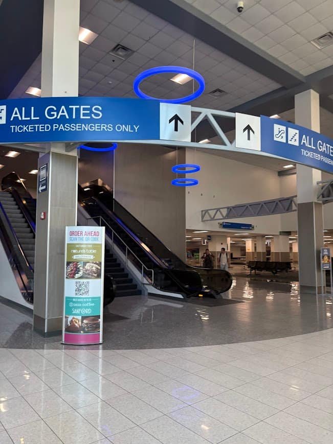 The main security entrance at Orlando Sanford International Airport with a blue "All Gates" sign and escalators. This illustrates the simple layout for travelers looking to fly to Florida on a budget through SFB.