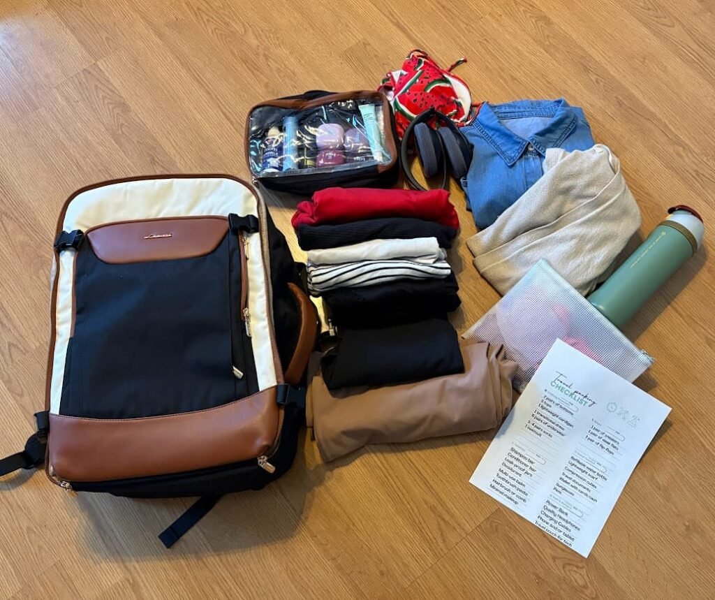 The LOVEVOOK 40L travel backpack laid out on a wood floor next to beach essentials like a swimsuit, folded clothes, a toiletry bag, and a travel packing checklist.