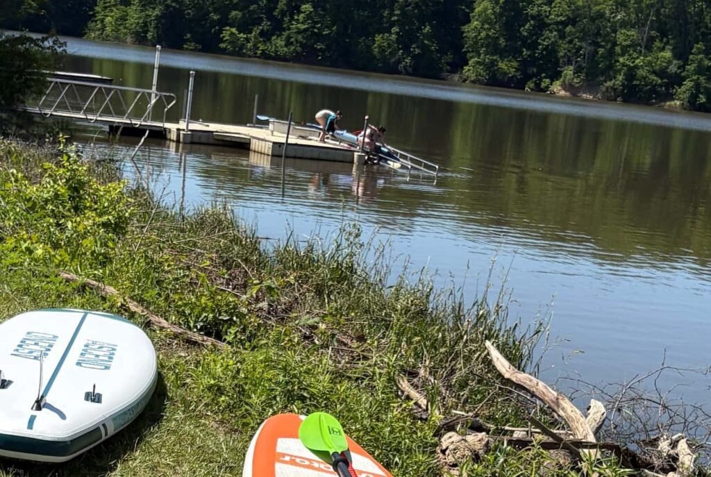 Two people preparing gear on a floating wooden dock launch platform with metal handrails on a calm, tree-lined lake, with two paddleboards resting on the grassy shore in the foreground.