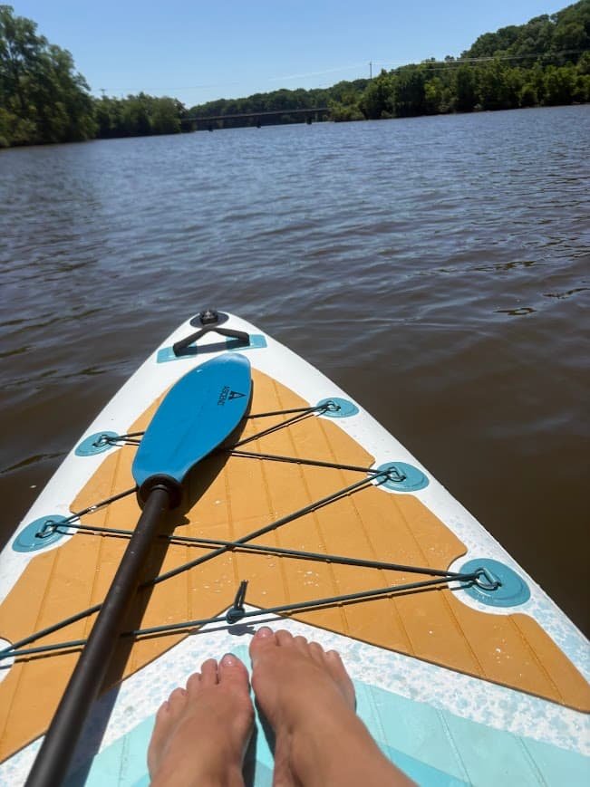 First-person view of bare feet on a blue and yellow stand-up paddleboard on a calm lake with a blue paddle across the deck.