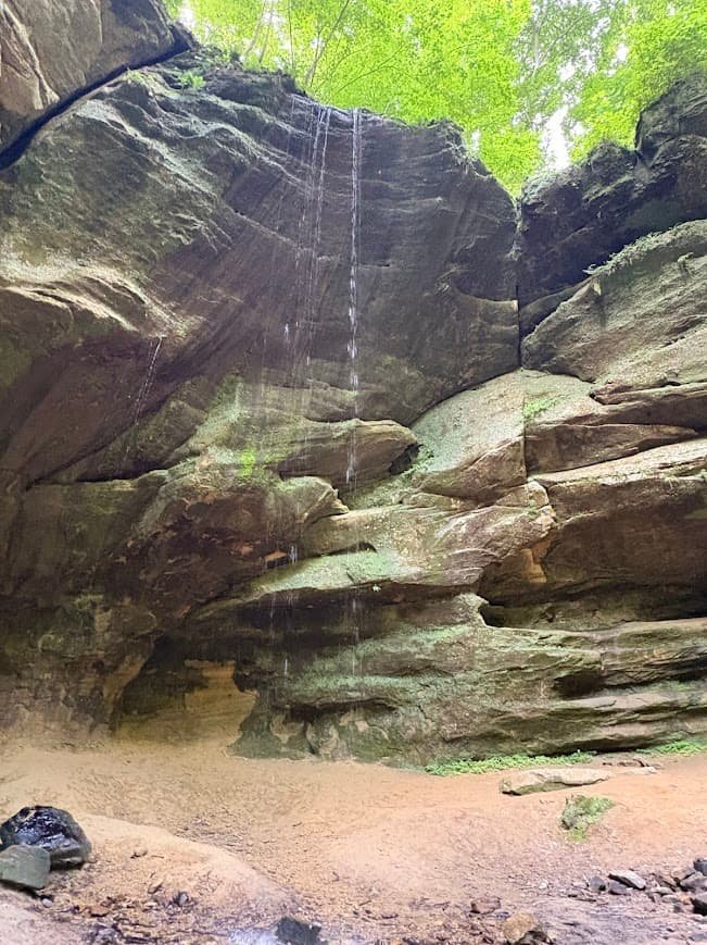 A high-angle sandstone overhang forms a deep recess cave where a delicate ribbon of water drips down onto a smooth, sandy floor.Planning a Mohican State Park hiking day trip?