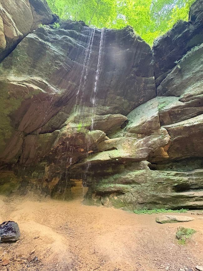 Planning a Mohican State Park hiking day trip? A gentle stream of water trickles over a high, curved sandstone rocky ledge into a sandy recessed alcove at Big Lyons Falls in Mohican State Park.