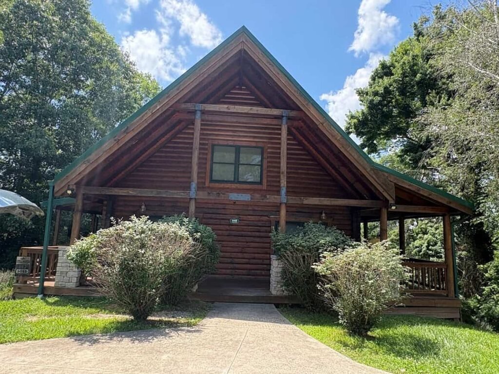 Front view of a rustic log cabin rental with an A-frame roof and covered porch, surrounded by green trees on a summer lake vacation.