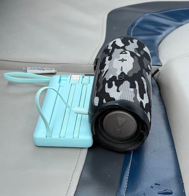 A light blue VRURC portable power bank charging a camouflage-patterned waterproof JBL Bluetooth speaker on a boat seat during a lake vacation.
