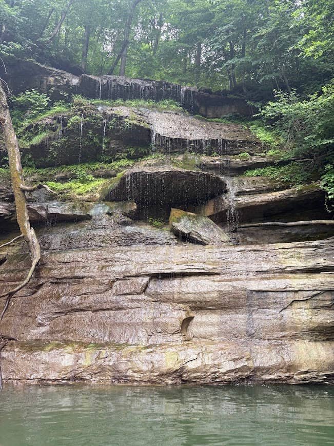 Water trickling down a tall, layered rock shelf waterfall surrounded by green trees into the lake during a lake vacation.