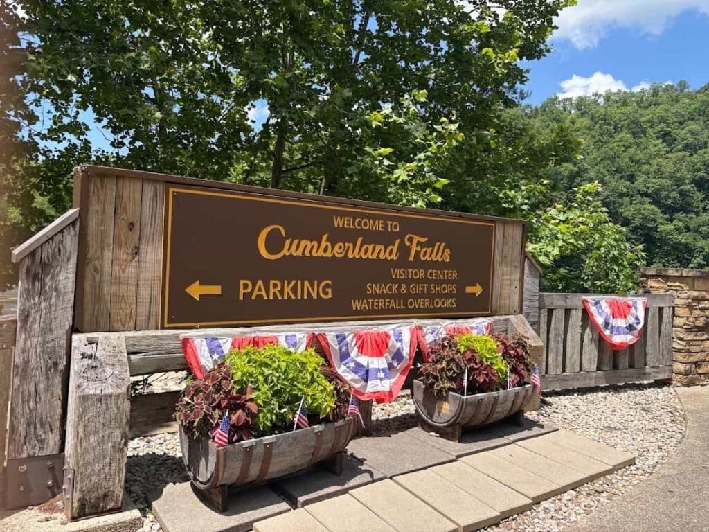 A large wooden welcome sign at Cumberland Falls State Resort Park that reads "Welcome to Cumberland Falls" with arrows directing visitors to parking, the visitor center, snack and gift shops, and waterfall overlooks.