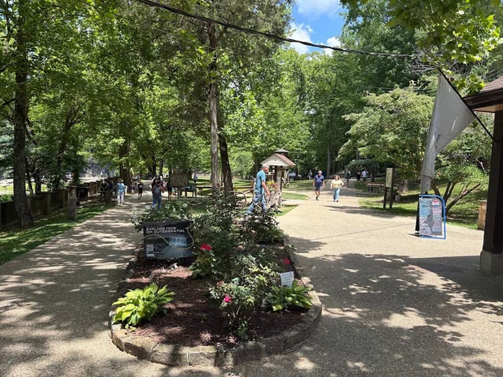 A wide paved walking path at Cumberland Falls State Resort Park, showing visitors walking along a shaded trail with green trees, flower beds, and park signage leading toward the visitor center.