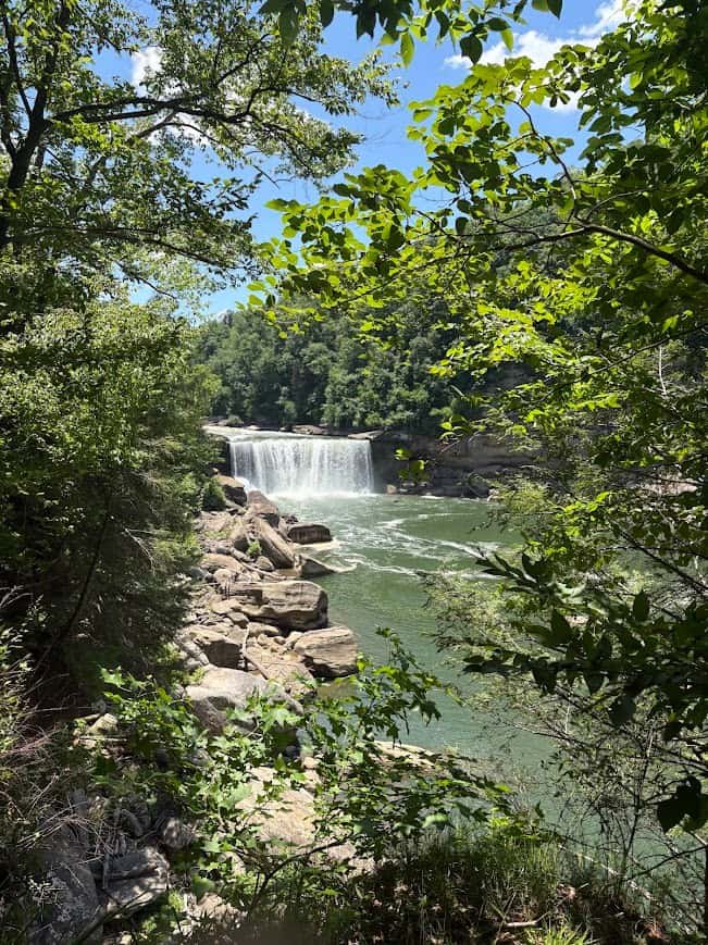 A scenic view of Cumberland Falls in Kentucky framed by green forest trees, showing the wide waterfall pouring into a river with large rocks lining the riverbank under a blue sky.