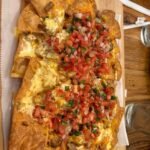 A top-down view of a large platter of loaded chicken nachos with melted cheese sauce and fresh tomato pico de gallo served on a wooden board at Charred Oak Grill. Title: charred-oak-grill-somerset-ky-chicken-nachos