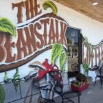 A colorful outdoor mural on a white brick wall that reads "The Beanstalk Coffee" with painted green vines and red-capped mushrooms, located next to black metal patio seating in downtown Somerset, Kentucky.