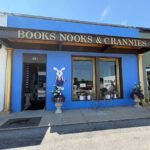 The bright blue brick storefront of Books Nooks & Crannies bookstore in downtown Somerset, Kentucky, featuring a large brown awning, gold columns, and an open doorway next to a large window display.