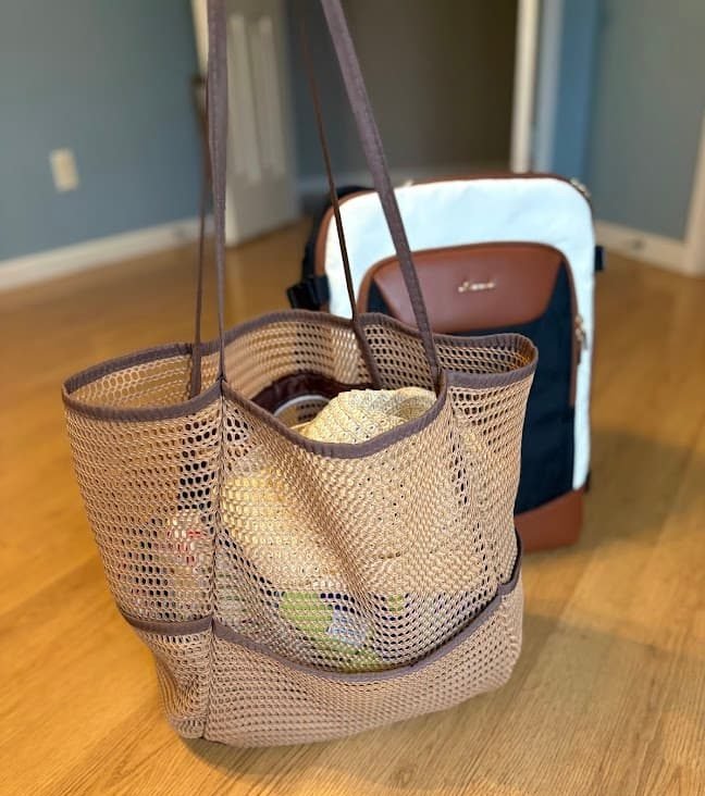 A tan mesh beach tote packed with summer essentials standing next to a white and brown travel backpack on a wooden floor for a lake vacation packing list.