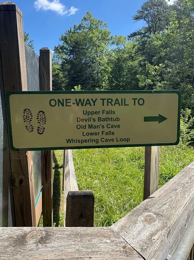 An official green and white park sign reading "One-Way Trail To Upper Falls, Devil's Bathtub, Old Man's Cave, Lower Falls, Whispering Cave Loop" along a wooden boardwalk at Hocking Hills State Park.