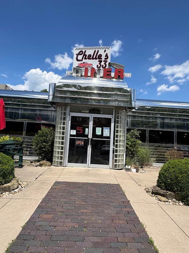 The retro, chrome exterior entrance of Chelle's 33 Diner on a bright, sunny day with a brick walkway leading to the glass front doors.