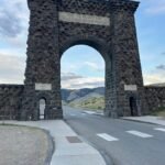Gardiner Montana north entrance Yellowstone Roosevelt Arch with mountains in the background during summer. Plan a Yellowstone Trip from Gardiner