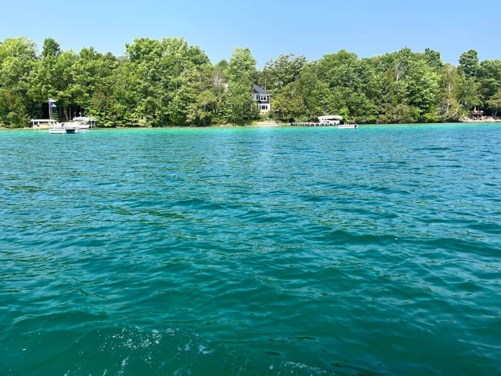 Wide view of clear, vibrant turquoise water at Torch Lake Michigan under a blue sky, featuring a lush green tree-lined shoreline with private docks and houses.