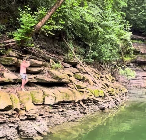 A person standing on a rocky, layered limestone shoreline fishing in the clear green water during a lake vacation.