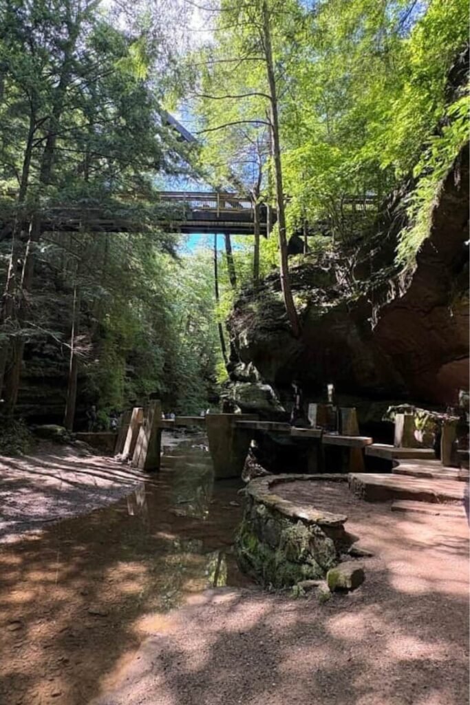 A scenic view inside a forested rock gorge at Hocking Hills State Park, featuring a high wooden footbridge overhead and concrete stepping stones crossing a shallow creek below.