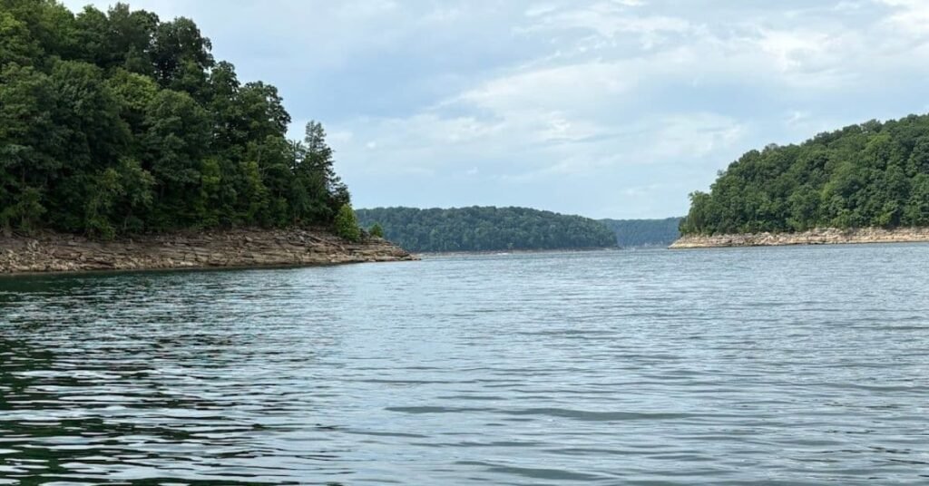 A wide view of the calm blue waters on a Lake Cumberland itinerary boat day, surrounded by lush green forested shorelines and rocky banks under a cloudy sky.