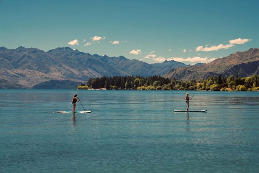 Two women standing up and paddling on wide boards across a vast, calm blue lake with massive mountains and pine trees in the background under a sunny sky.