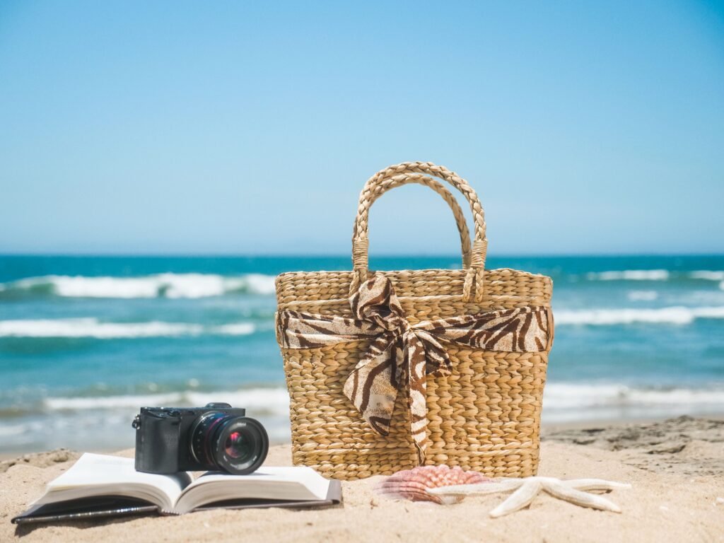 A serene beach scene with a woven bag, book, camera, and seashells, perfect for a relaxing day by the ocean.