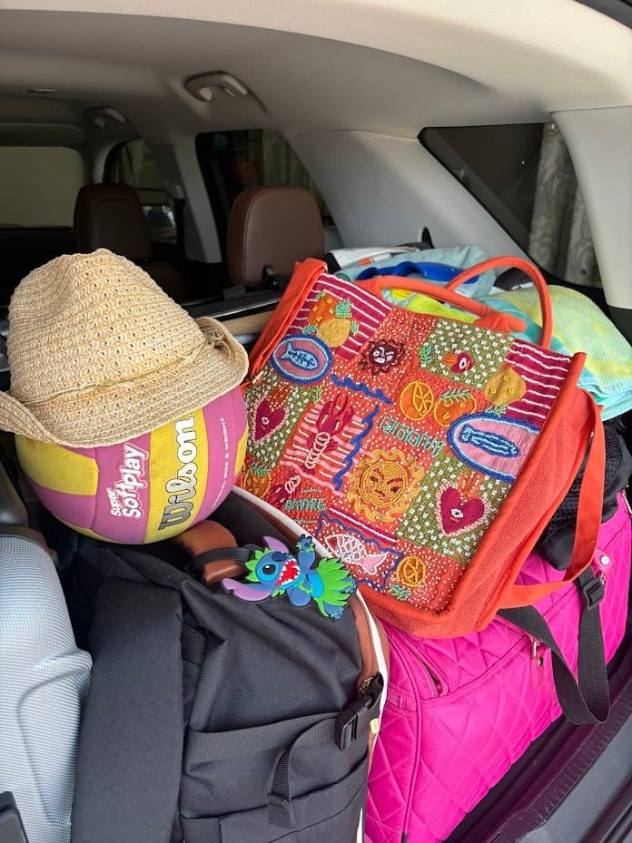 A packed car trunk filled with beach vacation luggage, featuring a vibrant orange hand-beaded tote bag with tropical motifs, a straw sun hat resting on a Wilson volleyball, a Stitch bag tag, and colorful luggage.