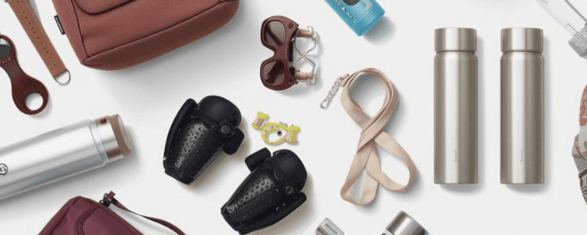 A collection of organized carry-on travel essentials displayed on a white background, including backpacks, a crossbody bag, water bottles, shoes, a scarf, headphones, and sunglasses, representing minimalist and intentional packing.